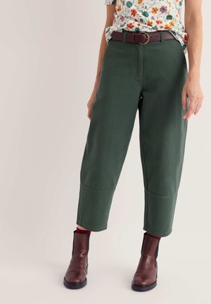 Personne portant un pantalon vert forêt court, une chemise à fleurs, une ceinture marron et des bottines marron foncé, debout devant un fond uni.