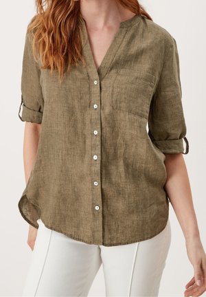 Kobieta w oliwkowozielonej lnianej koszuli na guziki z podwiniętymi rękawami do połowy przedramienia oraz białych spodniach, stojąca na jednolitym tle.