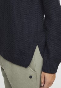 Pull en tricot bleu marine avec motif texturé, ourlet côtelé et fentes sur les côtés ; présente un bouton d'accent noir sur le côté inférieur.