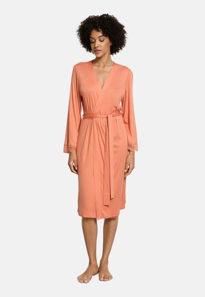 Kvinde stående barfodet iført en knælængde ferskenfarvet slå-om-kjole med lange ærmer og et bindebælte i taljen på en hvid baggrund.