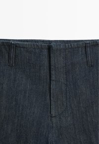 Dunkelblaue Denim-Hose mit Bund und Gürtelschlaufen sowie verdecktem Reißverschluss vorne, dargestellt vor weißem Hintergrund.