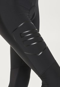Zwarte sportleggings van een glad, rekbaar materiaal, met reflecterende zwarte accenten en een strak ontwerp op de dij.