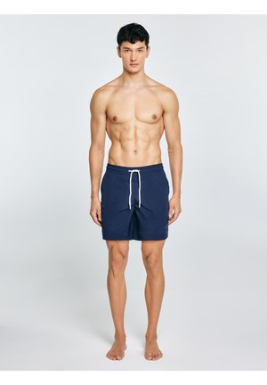 Młody mężczyzna o krótkich ciemnych włosach, stojący boso, ubrany w granatowe spodenki kąpielowe z białymwiązaniem na jednolitym jasnym tle.