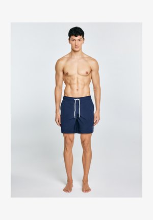 Młody mężczyzna o krótkich ciemnych włosach, stojący boso, ubrany w granatowe spodenki kąpielowe z białymwiązaniem na jednolitym jasnym tle.