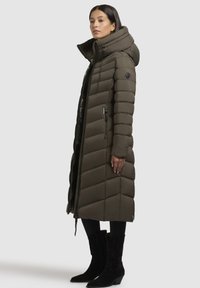 Lang, olivengrøn puffjakke med høj krave, quiltet mønster, lynlåslommer og sort metal, iført med tætsiddende sorte bukser og støvler.