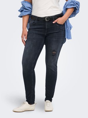 Person iført mørke skinny jeans med revet knæ, hvide sneakers, en hvid top stoppet ind og en blå stribet skjorte hængt over skuldrene.
