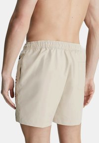 Costumi da bagno beige con vita elasticizzata, tasca sul retro e dettaglio della cucitura laterale. Realizzati in materiale leggero e morbido.