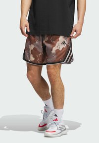 Herre brune camouflage shorts med lyse og mørke pletter, sorte sidestreger og en sort talje, iført hvide og røde sneakers.