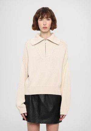 Beżowy sweter z dzianiny z dużym kołnierzem, zamkiem z przodu i ściąganym dołem, w zestawie z czarną skórzaną spódnicą mini. Luźny, oversize'owy krój.