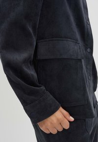 Giacca in corduroy nera con texture a coste, dotata di tasca a toppa sul petto e polsini con bottoni. Dettaglio ravvicinato della manica e della tasca.