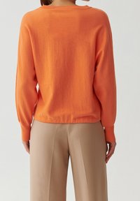 Pomarańczowy sweter o luźnym kroju, z prążkowanymi mankietami i ściągaczem, zestawiony z beżowymi szerokimi spodniami, ukazujący miękką fakturę i prosty design.