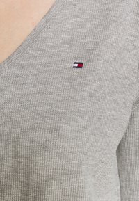 Grå strikket sweater med ribbet tekstur, der har en V-hals og et lille broderet logo i rød, hvid og blå på venstre bryst.