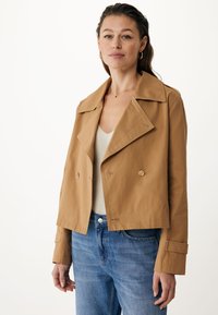 Giacca cropped color beige realizzata in tessuto morbido, caratterizzata da un design doppiopetto, un ampio colletto e discreti bottoni dorati. Indossata sopra un top chiaro.