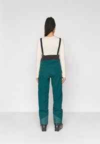 Τιρκουάζ χιονοστολές με μαύρες τιράντες και τσέπες με φερμουάρ, κατασκευασμένες από ανθεκτικό υλικό, διαθέτοντας χαλαρή εφαρμογή και τελειώματα με στενές γάμπες.