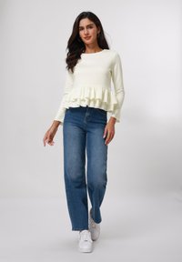 Camiseta blanca de manga larga con un dobladillo con volantes, combinada con jeans rectos de color azul claro y zapatillas blancas. Diseño sencillo sin patrones visibles.