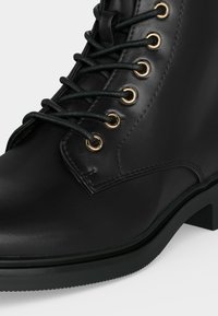 Bottine en cuir noir avec des lacets noirs passés à travers six paires d'œillets dorés, dotée d'un petit talon et de surpiqûres décoratives.