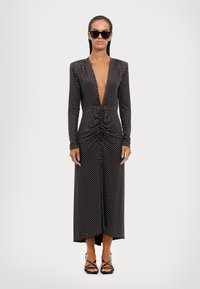 Zwarte lange mouw jurk met diepe V-hals, geplooid middel, witte stippen en enkellange zoom; gecombineerd met zwarte zonnebril en sandalen met bandjes.