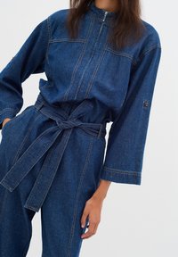 Denim-Overall mit hohem Kragen, Frontreißverschluss, weiten Ärmeln und einem gebundenen Gürtel. Verfügt über Kontrastnähte und eine entspannte Passform.