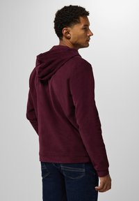 Burgundy hoodie i en texturerad, mjuk tyg och med en känguruficka. Modellen bär mörkblå jeans.