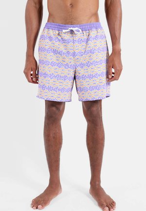 Homme portant un short de bain violet avec un motif à rayures ondulées orange, blanches et bleues, debout pieds nus sur un fond uni.