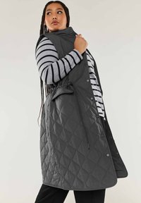 Gilet lungo trapuntato in grigio scuro, caratterizzato da un alta colletto, bottoni a pressione, tasche laterali e coulisse strategica in vita per una regolazione ottimale.