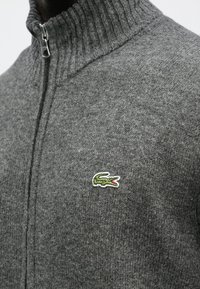 Sivý pletený sveter na zapínanie na zips s rebrovaným golierom a zeleným logom krokodíla na hrudi. Vyrobený z jemnej textúrovanej zmesi vlny.