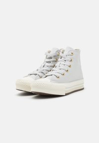 Zapatillas altas de lona gris claro con punteras de goma blanca, paneles laterales texturizados, ojales dorados y un diseño de suela en varias capas.