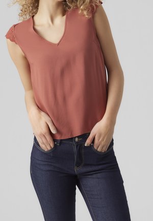 Vrouw met krullend haar, gekleed in een mouwloos roestkleurig topje en donkerblauwe spijkerbroek, met handen in de voorzakken tegen een effen grijze achtergrond.