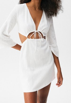 Femme portant une mini robe blanche texturée avec un décolleté en V profond, une découpe frontale nouée avec des cordons et des manches trois-quarts.