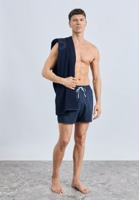 Tamsiai mėlyni plaukimo šortai su elastine juosmens juosta ir raišteliu. Basas modelis laiko tamsų rankšluostį ant vieno peties, demonstruodamas lygią odos tekstūrą.