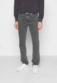 Grå denimjeans med rak passform, femficksdesign och metallknappsstängning, kombinerade med en svart långärmad tröja och vita sneakers.