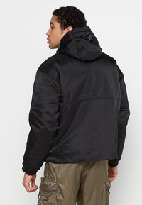 Svart vadderad jacka med huva, med en slät yta och elastiska muddar. Designen inkluderar en söm över ryggen och minimal detaljering.