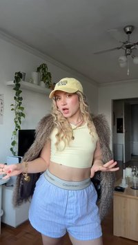 Chapéu amarelo com texto, top curto amarelo claro, xale de pelo e calções listrados azul claro com uma faixa cinza com a inscrição "Calvin Klein."