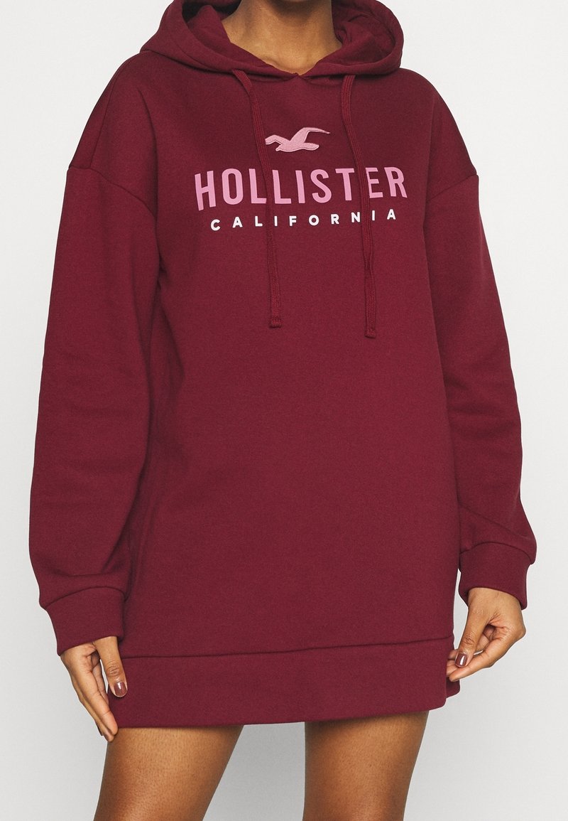 Sudadera con capucha de color burdeos y corte holgado, con un bolsillo canguro en la parte frontal, cordones y texto impreso "HOLLISTER CALIFORNIA".