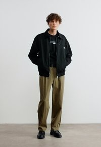 Νεαρός άνδρας που στέκεται με τα χέρια στις τσέπες, φορώντας μαύρο μπουφάν, μαύρο πουκάμισο με λευκό κείμενο, χακί παντελόνια και μαύρα παπούτσια μπροστά σε λευκό τοίχο.