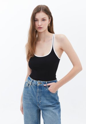 Zwart geribbelde tanktop met witte banden, gecombineerd met hooglopende blauwe spijkerbroek. Model heeft lang haar en poseert casual.