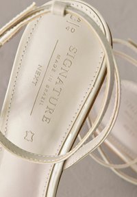 Sandali bianchi in pelle con cinturini sottili e una texture liscia. Presentano un marchio in rilievo e dettagli sulla taglia sulla soletta.