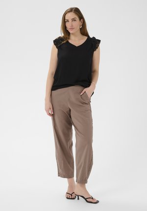 Kobieta stojąca, ubrana w czarny top bez rękawów z koronkową lamówką, taupeowe spodnie o długości 7/8 z ozdobnymi detalami po bokach oraz czarne sandały na obcasie.