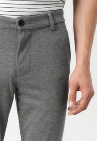 Gray trousers with a textured fabric, featuring a single button closure, belt loops, and side pockets. Close-up of hand near the waistband.