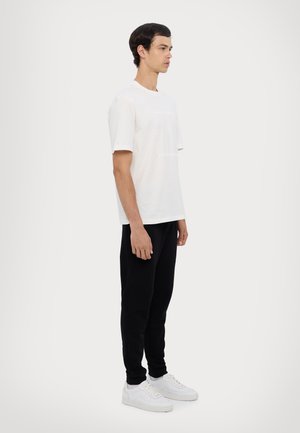 T-shirt bianca in cotone con maniche corte e testo grafico, abbinata a pantaloni jogger neri e sneaker bianche. Sfondo semplice.
