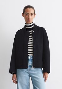 Sort bomberjakke med ribbet krave og frontknaplukning. Bæres over en sort og hvid stribet turtleneck, parret med lyseblå jeans.