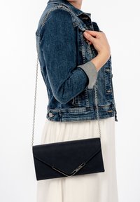 Pochette en similicuir noir avec une bandoulière chaîne argentée, dotée d'un rabat triangulaire et d’un détail de fermeture élégant, portée sur une robe blanche.