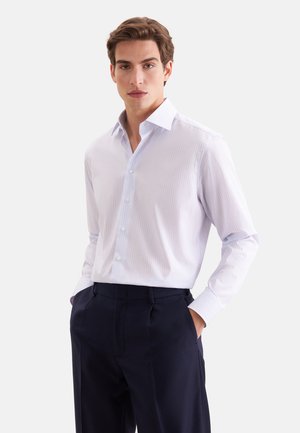 Jeune homme portant une chemise rayée bleu clair à boutons et un pantalon bleu marine foncé, les mains dans les poches, debout devant un fond uni.