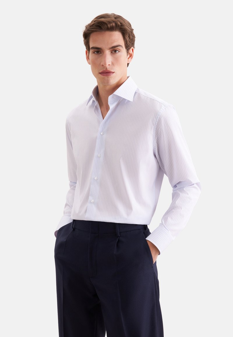 Jeune homme portant une chemise rayée bleu clair à boutons et un pantalon bleu marine foncé, les mains dans les poches, debout devant un fond uni.