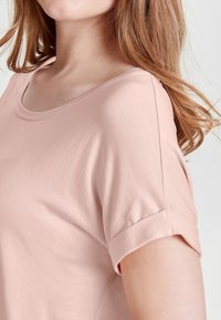 Helles rosa Kurzarmoberteil mit einem runden Ausschnitt, weichem Material und umgeschlagenen Ärmeln. Der Stoff wirkt glatt und leicht.