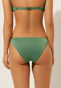Groene bikini broekje gemaakt van glanzend materiaal met een soepele textuur. Heeft een lage snit en minimale bedekking. Verstelbare zijkanten.
