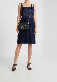 Abito blu a pieghe con scollatura a carré, spalline regolabili; borsa nera con lettering dorato; tacchi neri a punta con dettagli in raso.