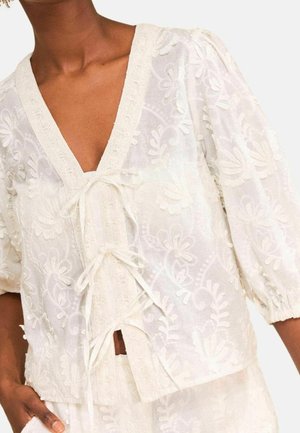 Mujer con blusa blanca bordada con flores, mangas abullonadas y lazos atados al frente, mostrada desde el cuello hasta las caderas.