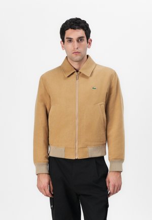Tan zip-up jacket with a collar and ribbed hem. Features side pockets and a small green logo. Fabric has a smooth, soft texture.
