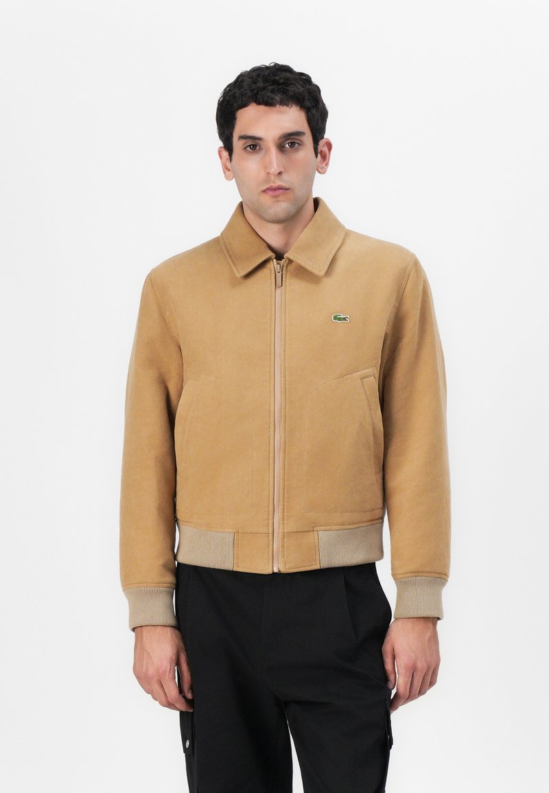 Veste zippée beige avec col et ourlet côtelé. Dispose de poches latérales et d'un petit logo vert. Le tissu a une texture lisse et douce.